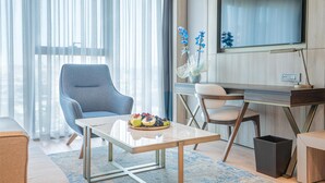 Une chambre d’hôtel moderne dotée d’une grande fenêtre, d’un téléviseur à écran plat fixé au mur, d’une table à manger avec un assortiment de fruits, d’un fauteuil bleu et d’un bureau en bois.