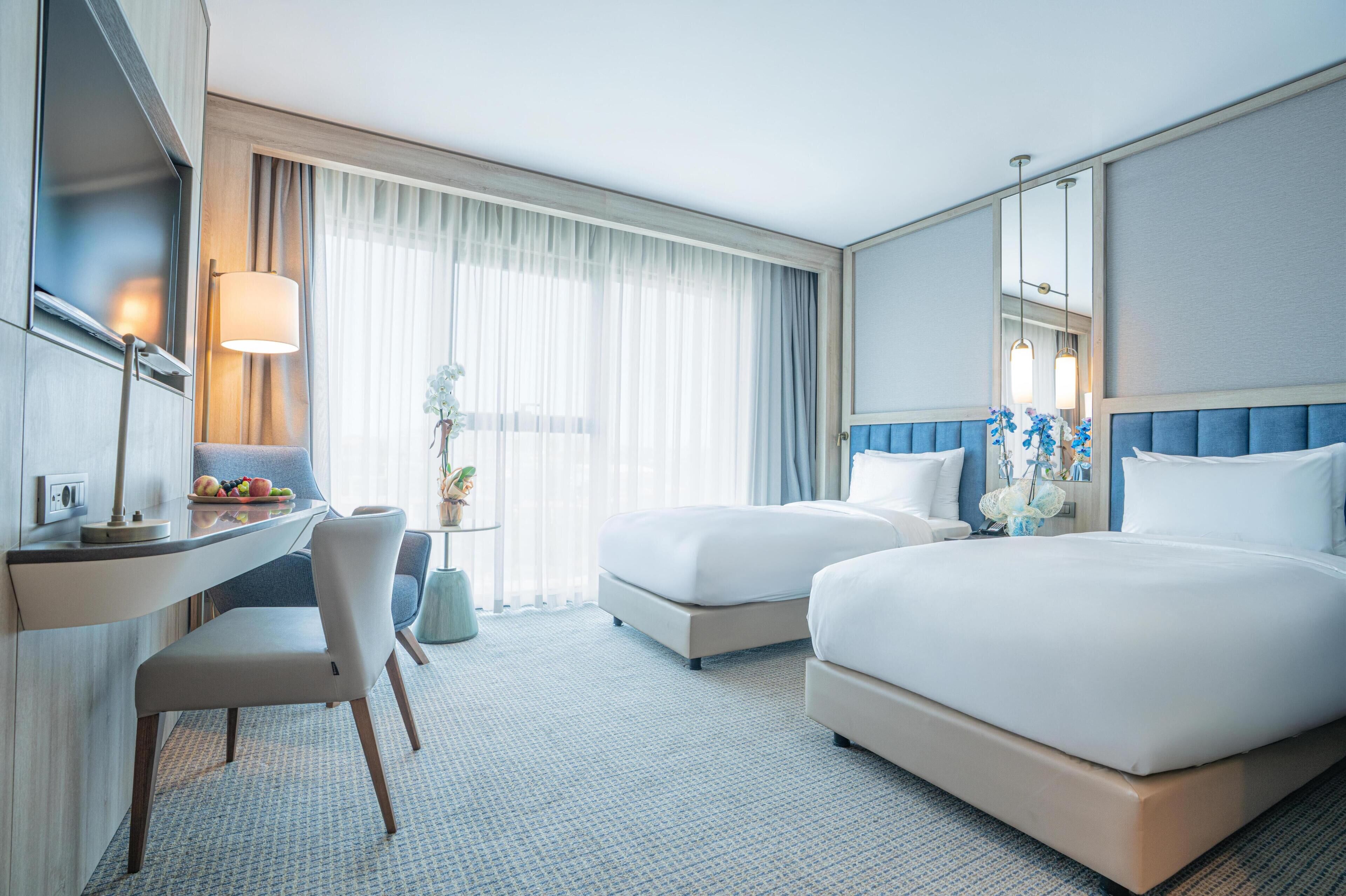 A hotel room with two beds, a desk with a chair, a small table with fruit, and a large window with curtains.