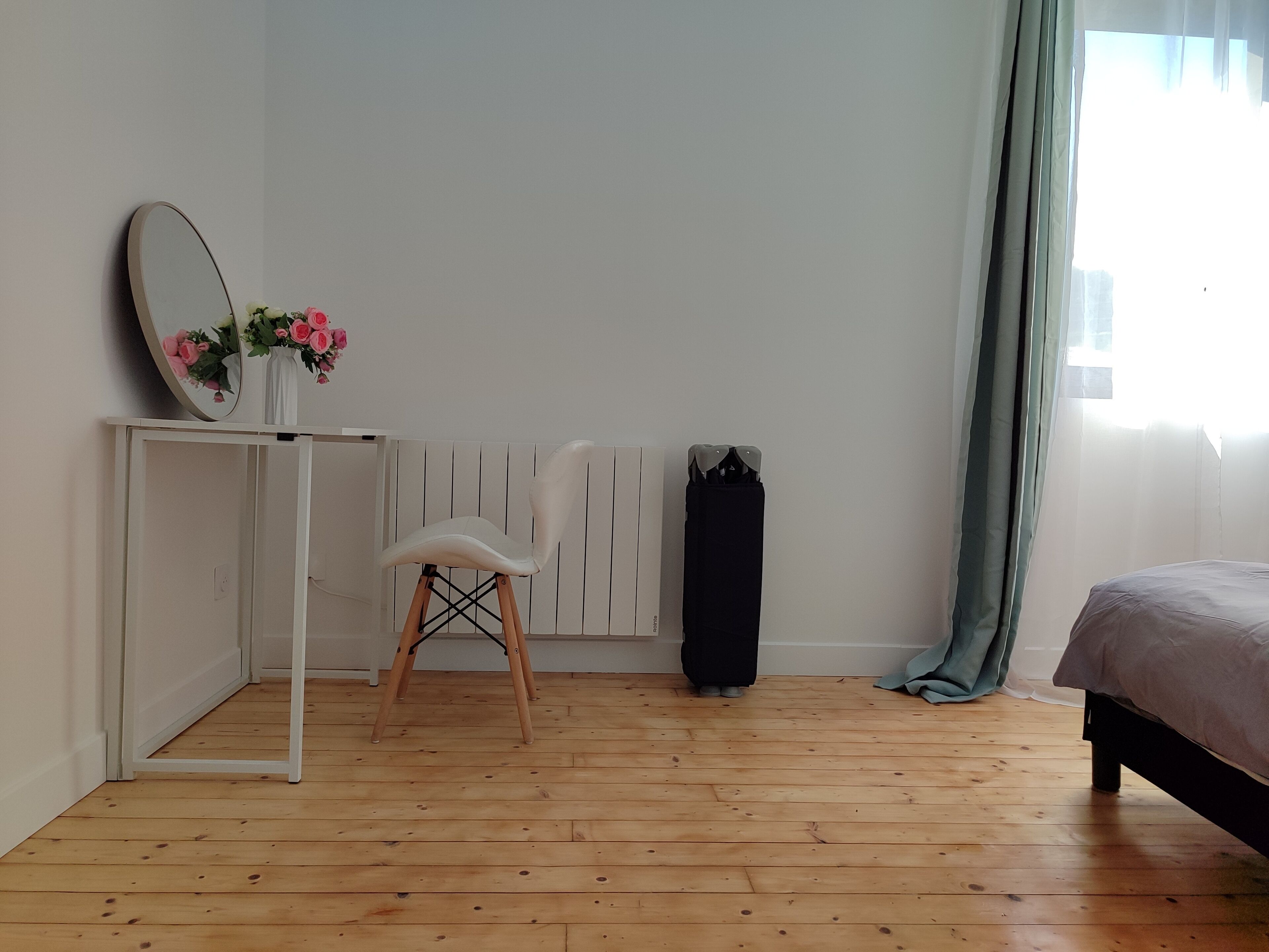Et værelse med trægulv, et hvidt skrivebord med spejl og en vase med blomster, en hvid stol, en radiator, en sort taske og en seng med gråt sengetæppe.