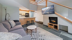 Una sala de estar moderna con un sofá, una mesa de centro, una chimenea, un televisor montado en la pared y una cocina al fondo.