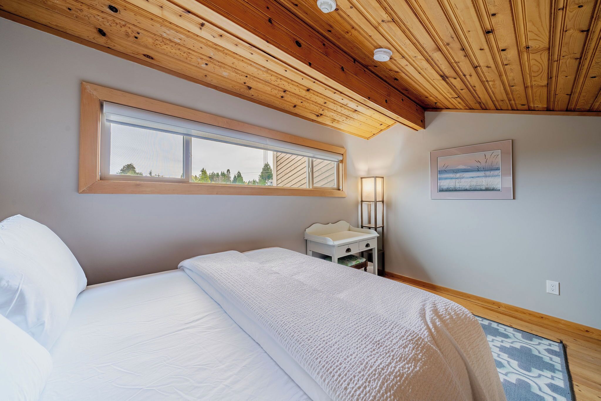 A bedroom with a wooden ceiling, a large window, a bedside table with a lamp, and a framed picture on the wall.