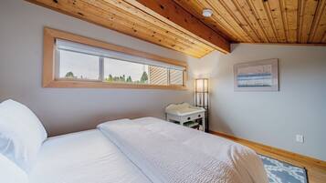 A bedroom with a wooden ceiling, a large window, a bedside table with a lamp, and a framed picture on the wall.