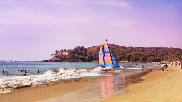 Una spiaggia con una barca a vela, persone che nuotano e una collina sullo sfondo.