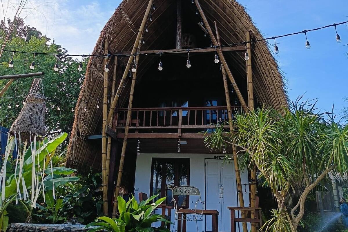 A thatched-roof building with a balcony and string lights, surrounded by lush greenery.
