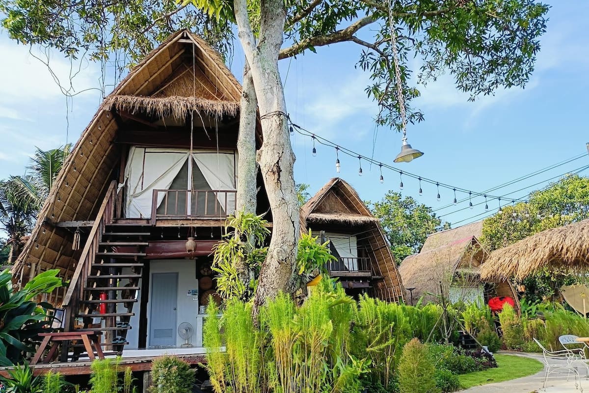 Two thatched-roof cottages with balconies, surrounded by lush greenery and a clear sky.