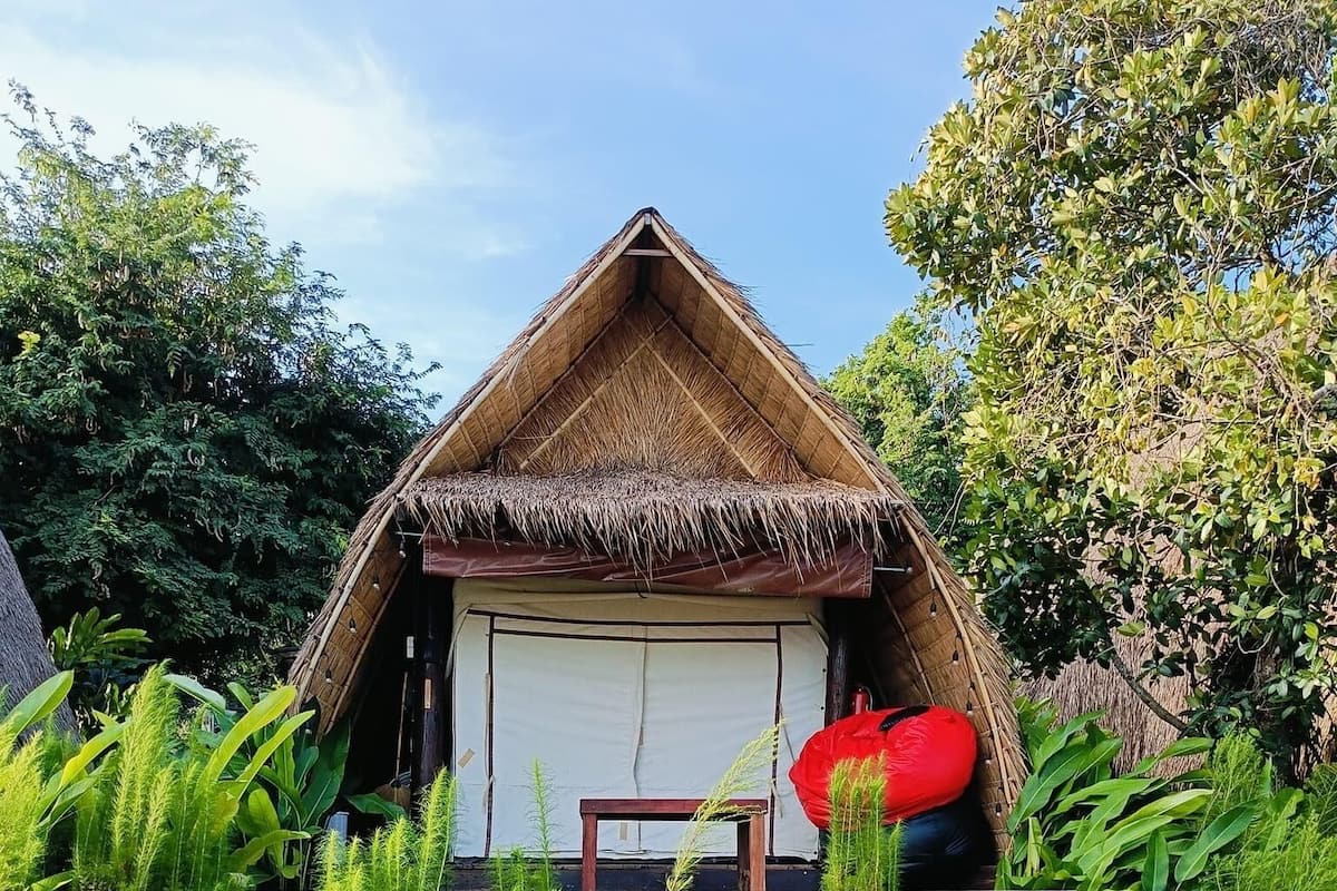 A thatched-roof structure with a bed and a table, surrounded by lush greenery and colorful flowers.