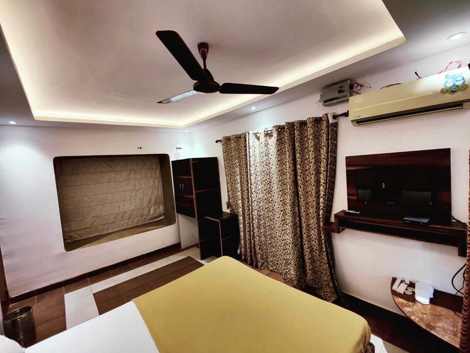 A modern hotel room with a bed, a ceiling fan, an air conditioning unit, a TV, and patterned curtains.