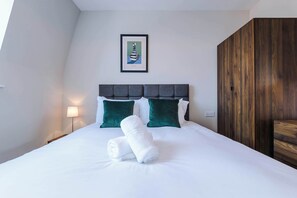 Un lit bien fait, avec du linge de lit blanc et deux serviettes enroulées, une lampe de chevet et une œuvre d’art encadrée au mur.