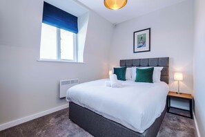 A neatly made bed with white linens and green pillows, a bedside table with a lamp, a framed artwork on the wall, and a window with blue blinds.