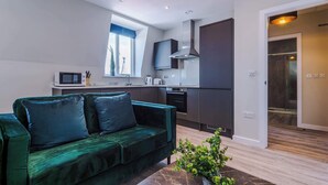 A modern living room with a green sofa, a kitchen area with dark cabinets, and a large window. - Elysée Suites - Chic 1 Bed Flat in Southport (Southport)