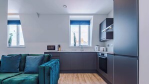 A modern kitchen with dark cabinetry, a built-in oven, and a microwave. There is a teal sofa with cushions in the living area. - Elysée Suites - Chic 1 Bed Flat in Southport (Southport)
