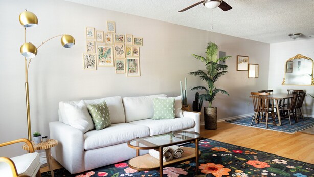 A living room with a white sofa, a glass coffee table, a floor lamp, and a large floral rug. There are framed pictures on the wall and a cactus plant.