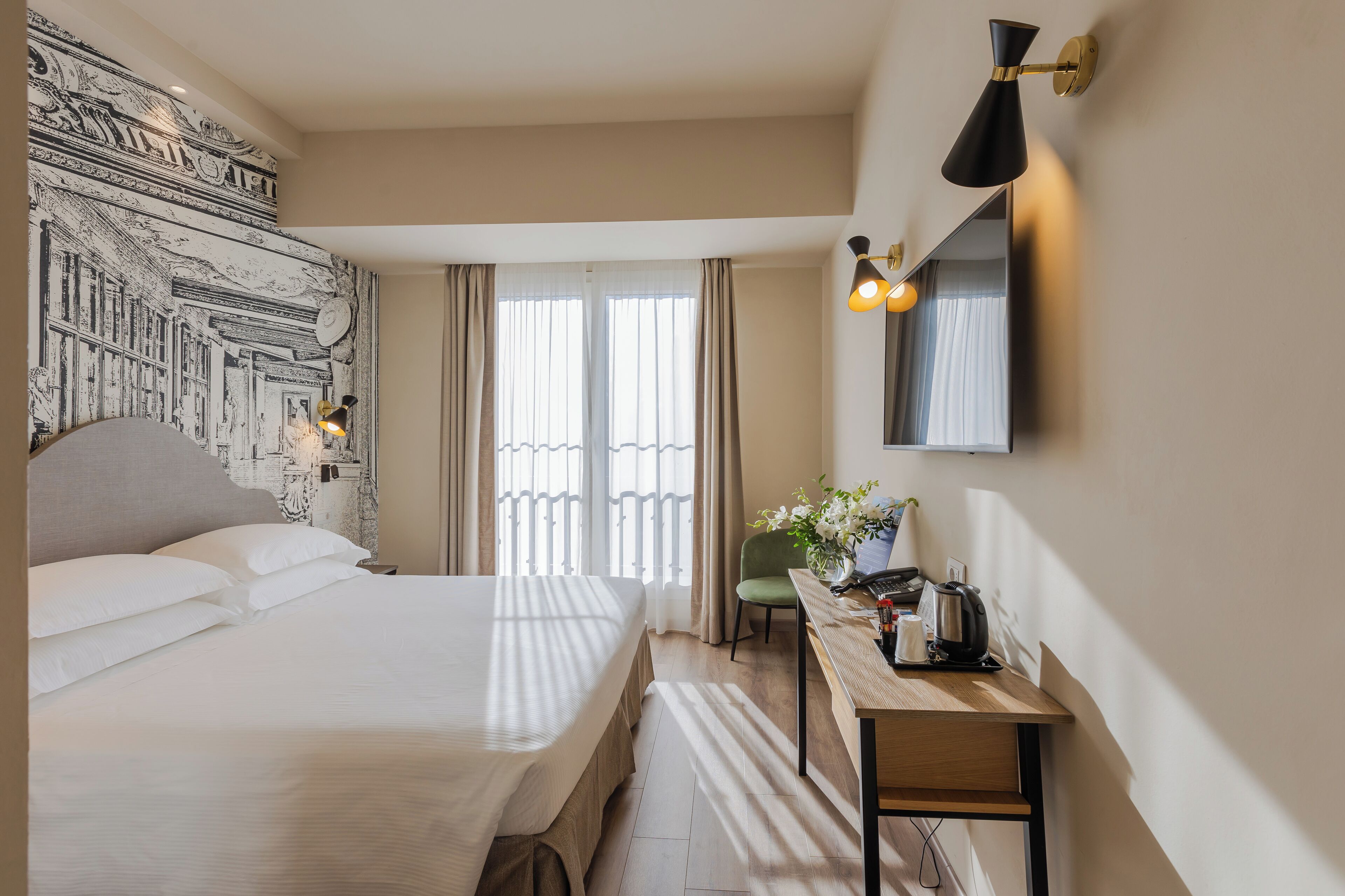 Une chambre d’hôtel avec un grand lit, un bureau avec une chaise, une lampe fixée au mur et une grande fenêtre avec des rideaux.