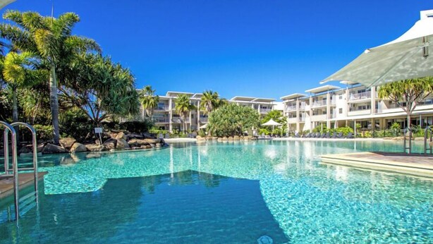 Piscina azul límpida rodeada por palmeiras e um prédio de resort com várias varandas.