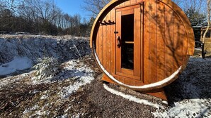 Une structure en bois en forme de tonneau, dotée d’une porte, qui se dresse sur un chemin de gravier parsemé de plaques de neige.