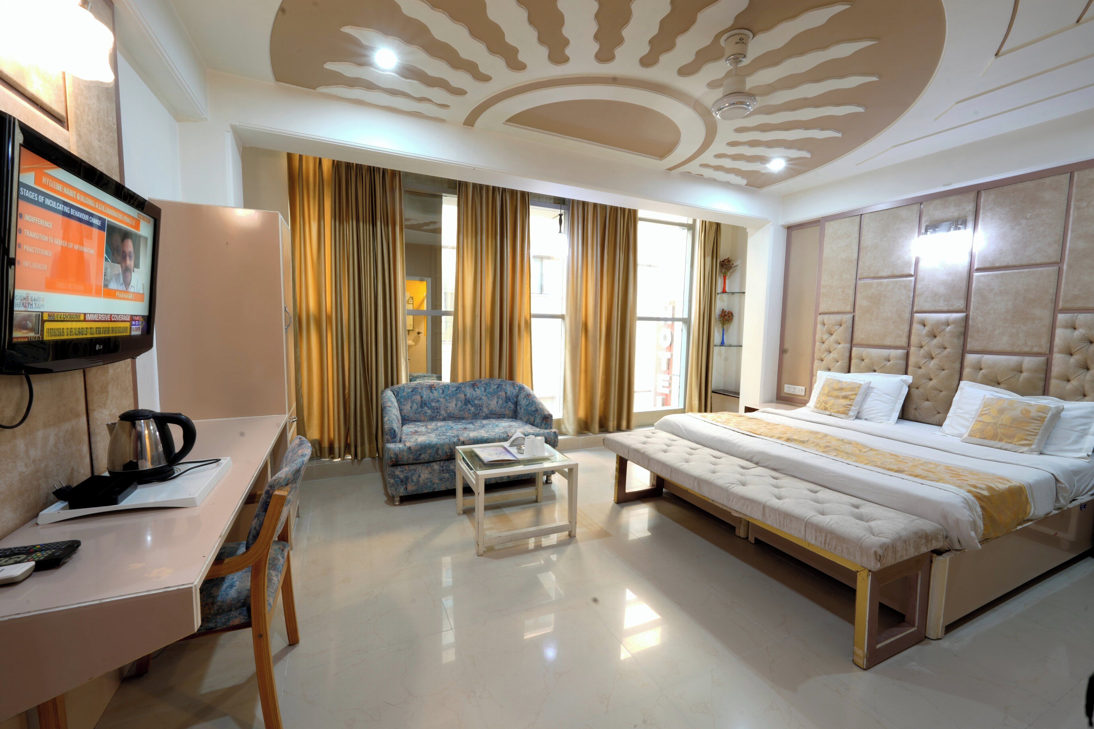 A hotel room with a bed, a sofa, a desk with a kettle, a television, and curtains.