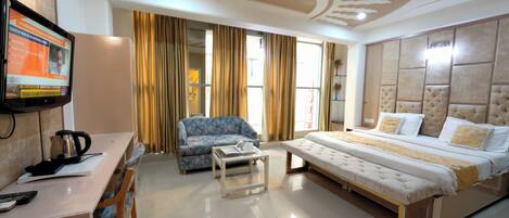 A hotel room with a bed, a sofa, a desk with a kettle, a television, and curtains.