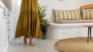 Une personne portant une robe de couleur moutarde traverse une pièce où se trouvent un canapé blanc, des coussins à rayures, un tapis tissé et une plante en pot ornée de fleurs rouges.