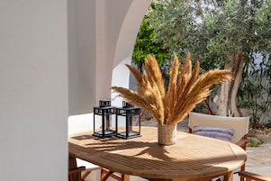 Una mesa de madera redonda con una cesta trenzada que contiene plumas de plumeros, dos faroles negros y un cojín en una silla blanca.