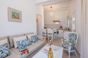Sala de estar con sofá, mesa de centro y adornos. Hay una zona de comedor con mesa y sillas. Se ve la cocina al fondo, con armarios blancos y una ventana.