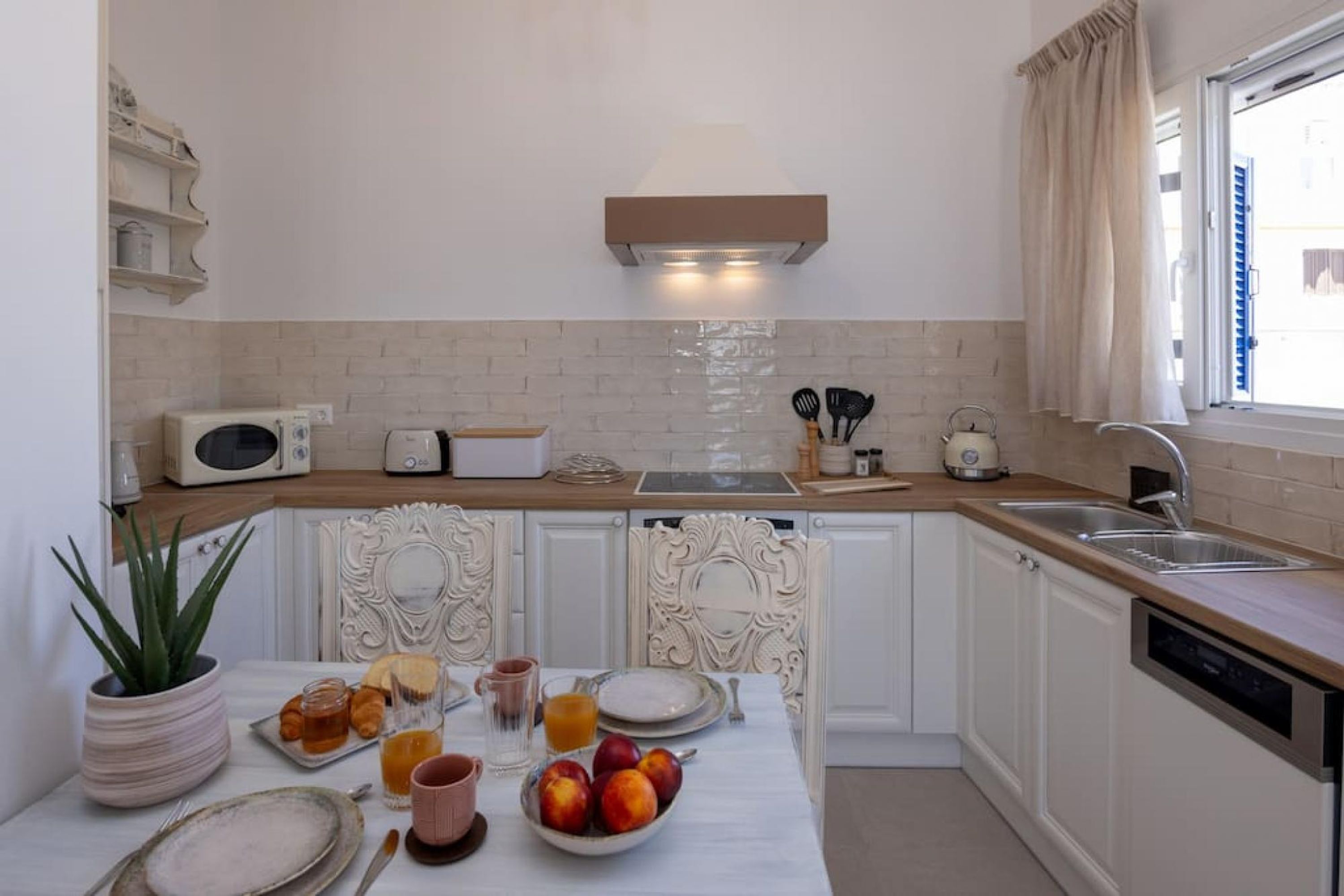 A compact kitchen with white cabinetry, a microwave, toaster, and a sink. There's a dining table with plates, glasses, and a fruit bowl.