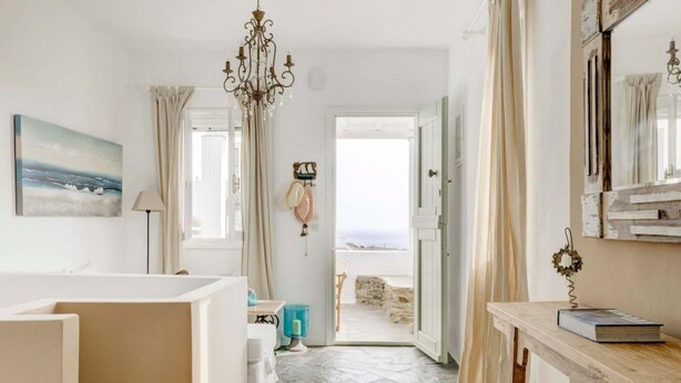 A room with a stone floor, a wooden table, a bed, a painting, and a view of the sea through an open door. - Ballos for 4 in Exomvourgo (Tinos)