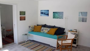 A room with a white sofa, a small side table, a chair, a standing fan, and a cart with items. - Amberi Tinos in Panormos (Tinos)