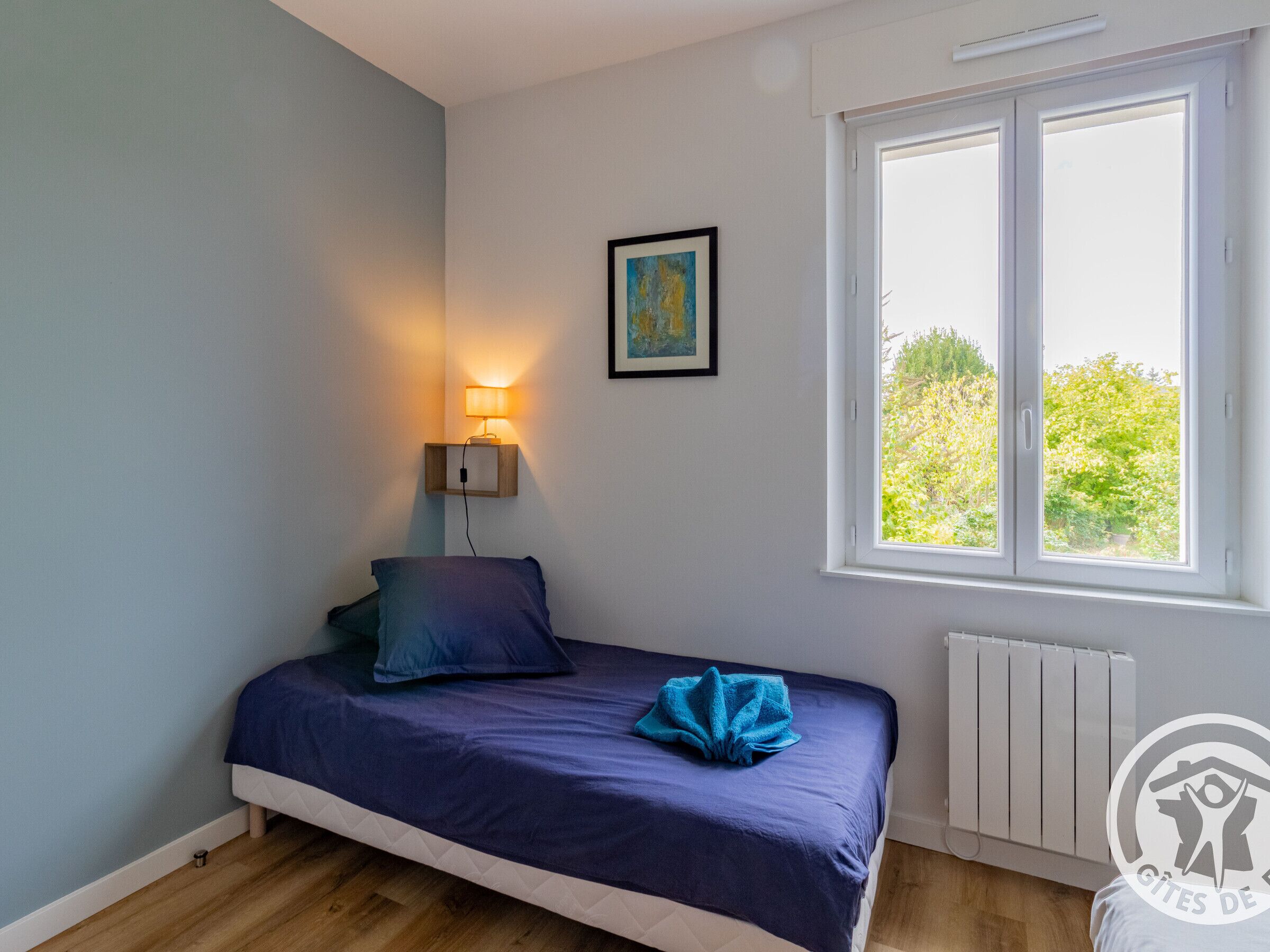 A single bed with a blue bedspread and pillows, a bedside lamp, a framed picture on the wall, and a window with a view of greenery.