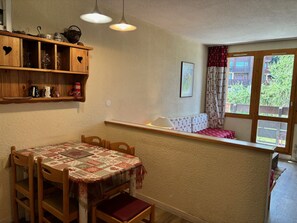 A cozy dining area with a wooden table and chairs, a shelf with kitchenware, and a window with a view of greenery.