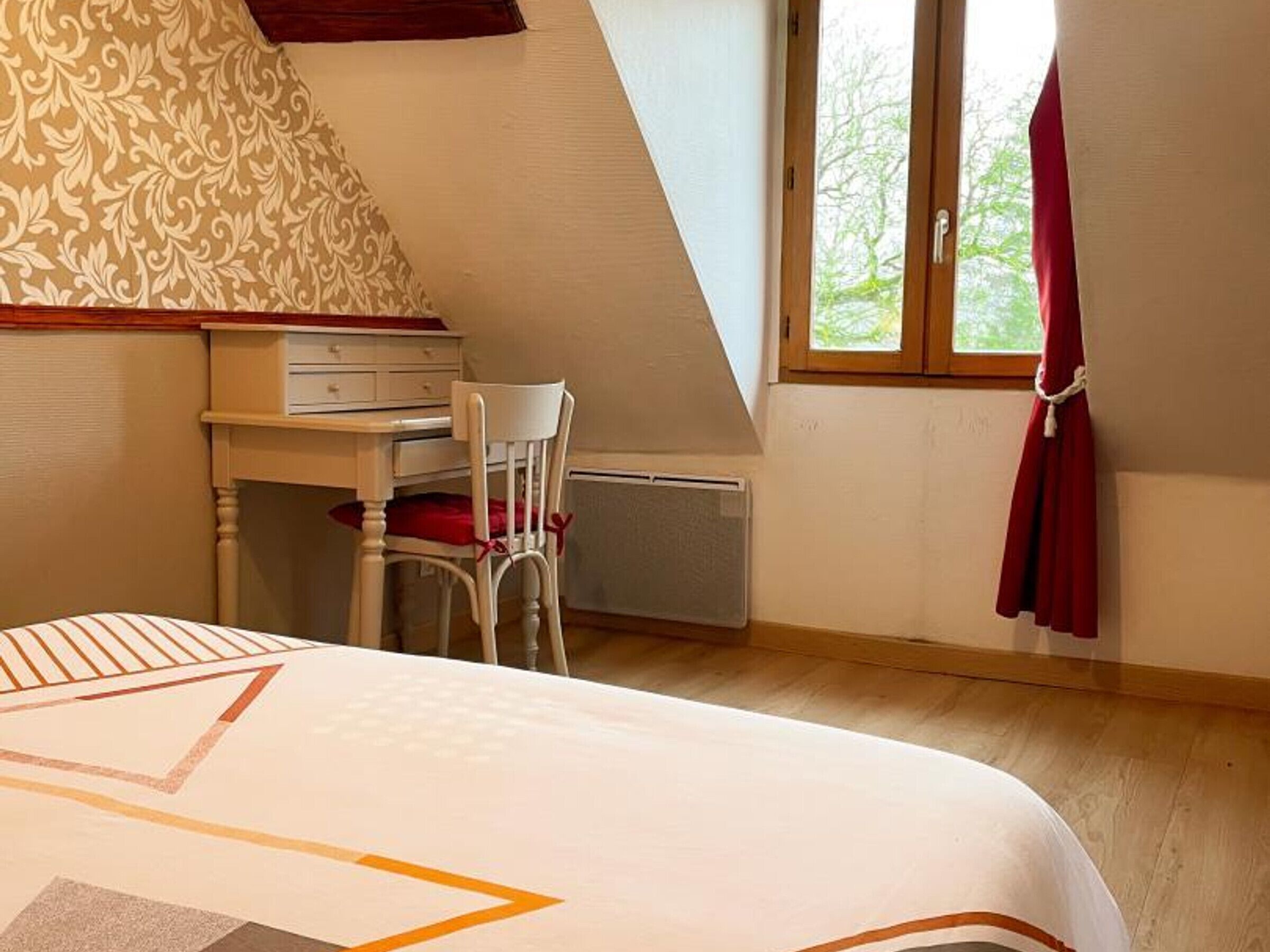 A bedroom with a bed, a desk, and a chair. There is a window with a view of trees.