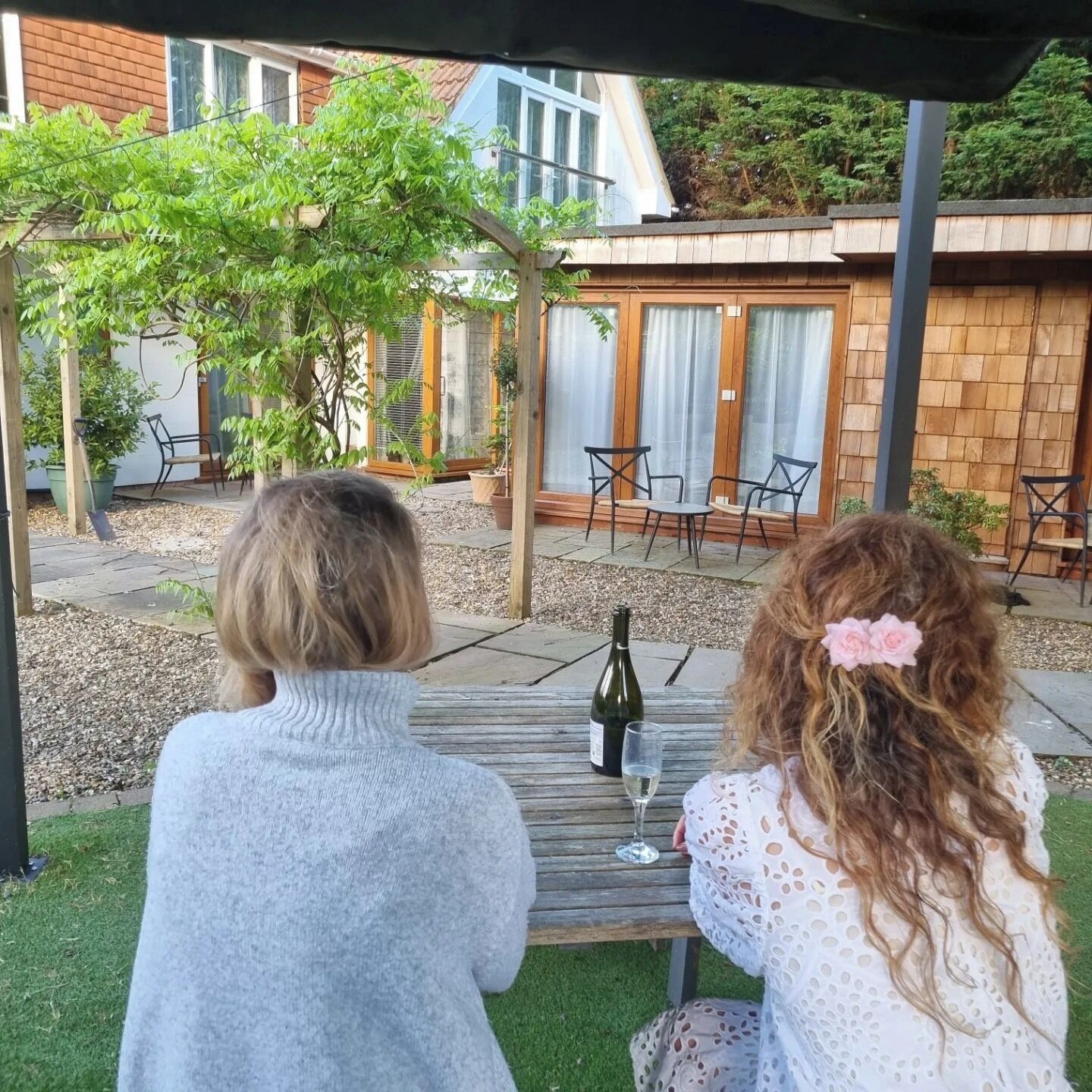 Zwei Personen sitzen an einem Tisch im Freien, mit einer Weinflasche und einem Glas, in einer Terrasse mit Stühlen und Blumentöpfen.