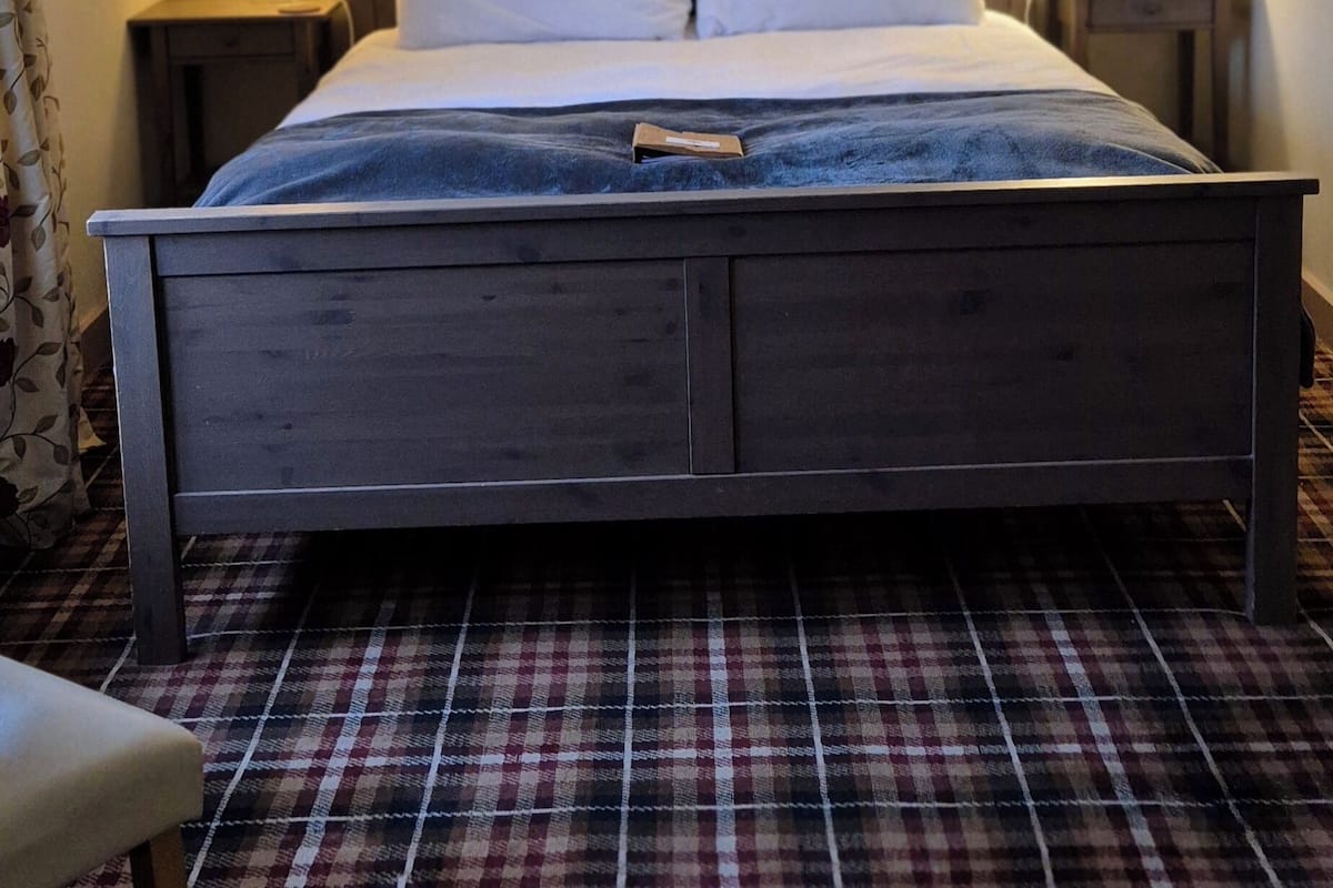 A neatly made bed with a wooden headboard, two bedside lamps, and a plaid-patterned carpet.