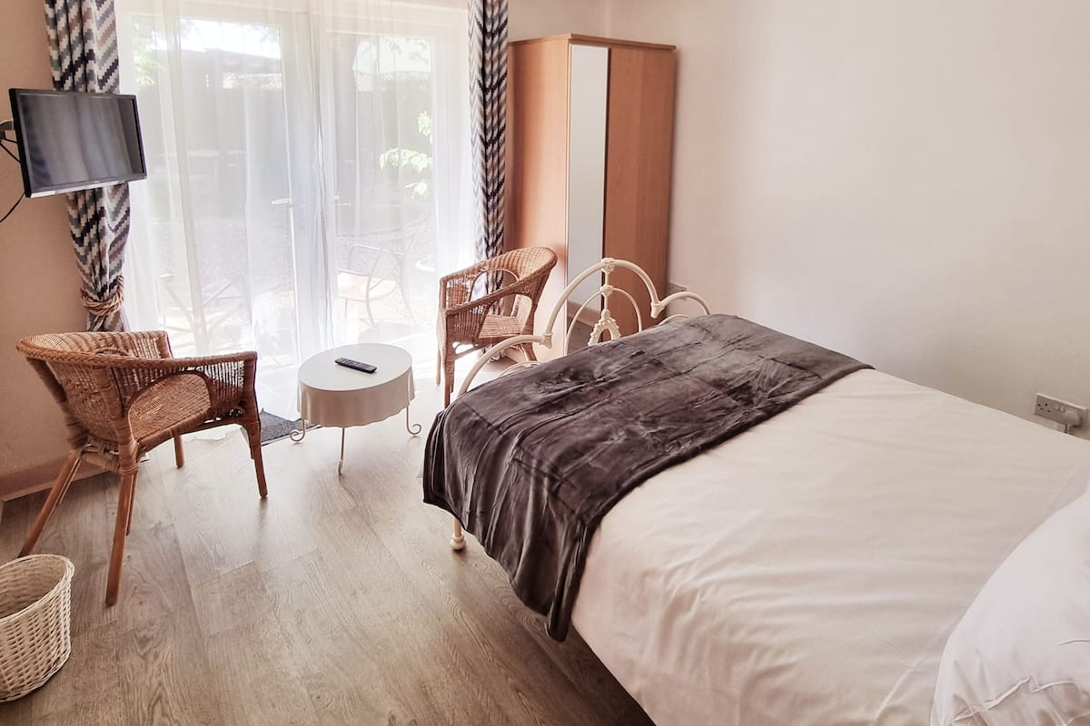A hotel room with a bed, two wicker chairs, a small round table, a TV, and a ceiling fan.