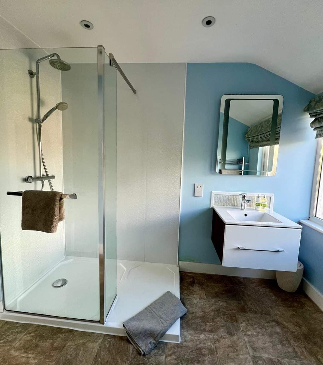 Ein modernes Badezimmer mit einer Glaskabine zur Dusche, einem weißen Waschtisch mit Spiegel und einem braunen Handtuch auf dem Boden.