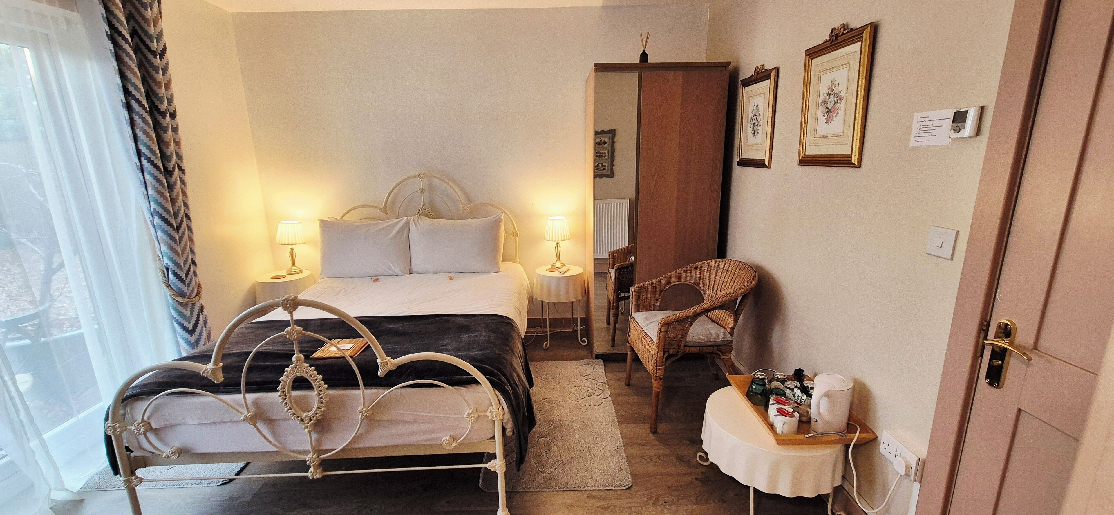 Un dormitorio con cama, lámparas de noche, una silla de mimbre, una mesita con artículos de tocador y una puerta con manija.