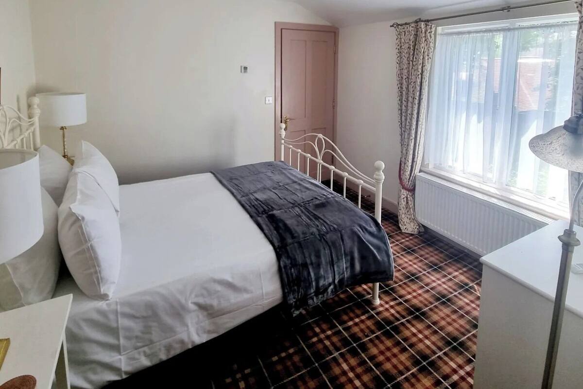 A hotel room with a bed, a nightstand, a lamp, a window with curtains, and a door.