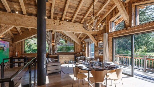 A spacious wooden interior with a high ceiling, a dining area with round tables and chairs, and large windows offering views of the outdoors.