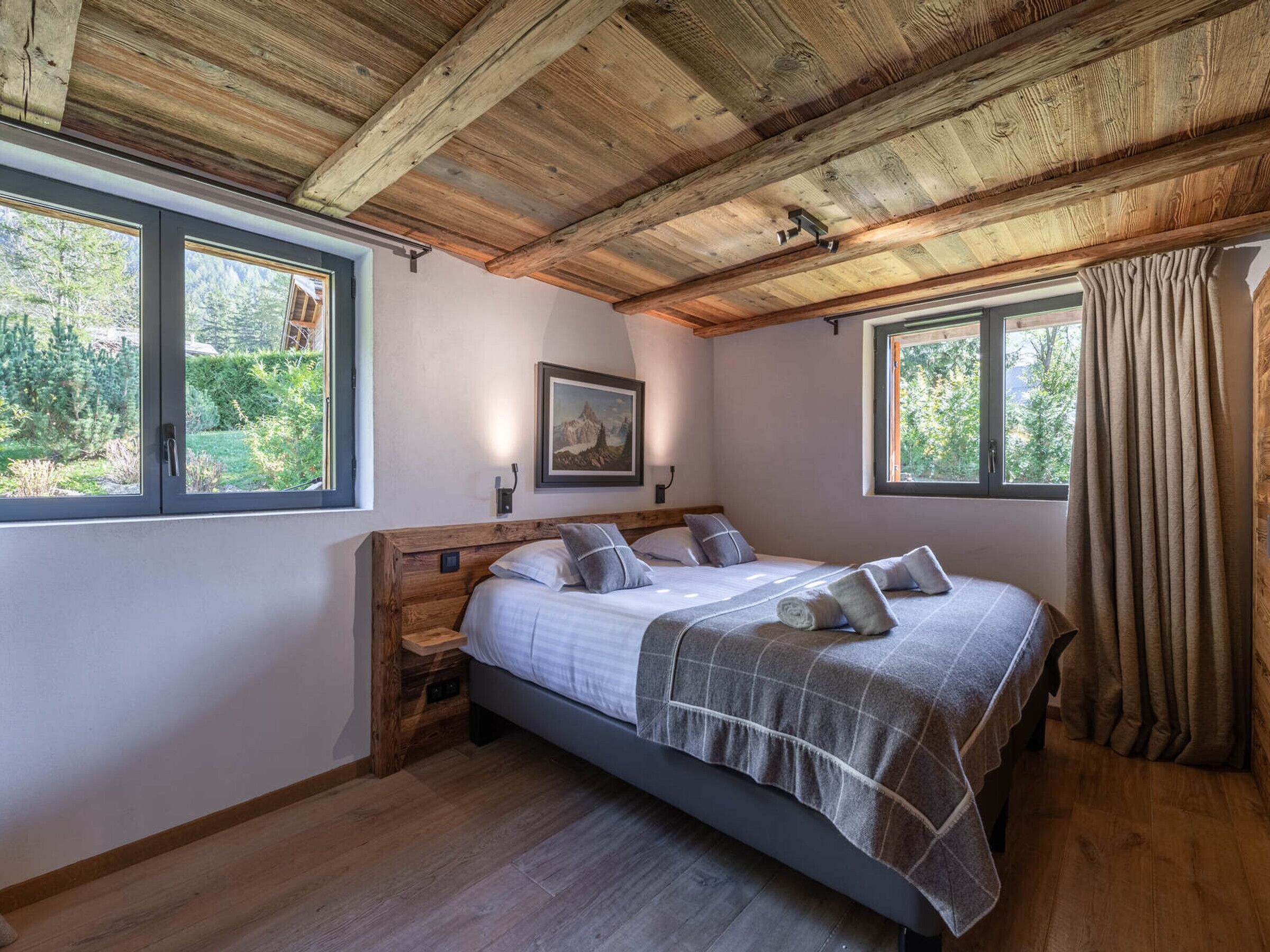 A bedroom with a wooden ceiling, two windows, a bed with a grey blanket, and a nightstand.