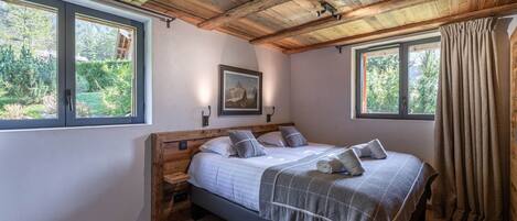 A bedroom with a wooden ceiling, two windows, a bed with a grey blanket, and a nightstand.