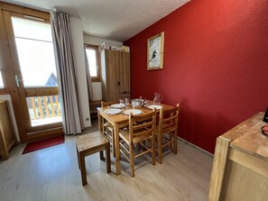A dining area with a wooden table set for four, a red wall, and a window with curtains. - Renovated 26sqm studio for 4 with balcony, ski-in/ski-out, pets allowed - Vallandry (Peisey-Vallandry)