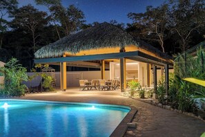 A poolside area with a covered seating area, a thatched-roof structure, and a clear night sky.