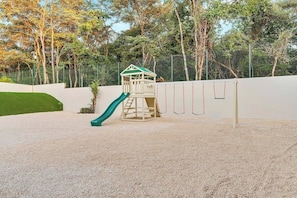 A playground with a slide and a climbing structure, surrounded by a fence and trees.