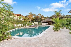 A swimming pool with a curved edge, surrounded by a stone patio, and a thatched-roof structure.