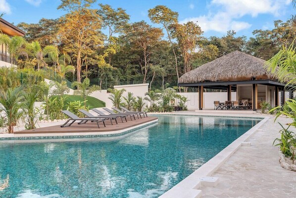 Una piscina con sillas de descanso, una estructura de techo de paja y follaje circundante.