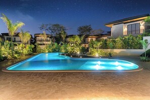 A swimming pool with a stone deck, surrounded by tropical plants and modern houses under a starry night sky.