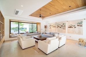Ein modernes Wohnzimmer mit großem Fenster, weißen Sofas, einer Holzbalkendecke und Wandkunst aus Makramee.