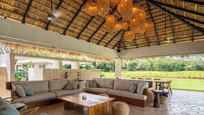 An outdoor lounge area with a thatched roof, stone flooring, and a mix of seating arrangements including sofas and chairs. - Seasons at Ceiba (Hacienda Pinilla)