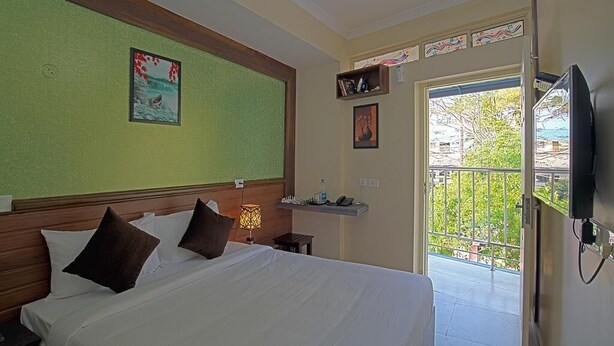 A hotel room with a bed, a nightstand, a wall-mounted TV, a window with a view of greenery, and a door leading to a balcony. - Gems9 Airport Hotel (Nedumbassery)