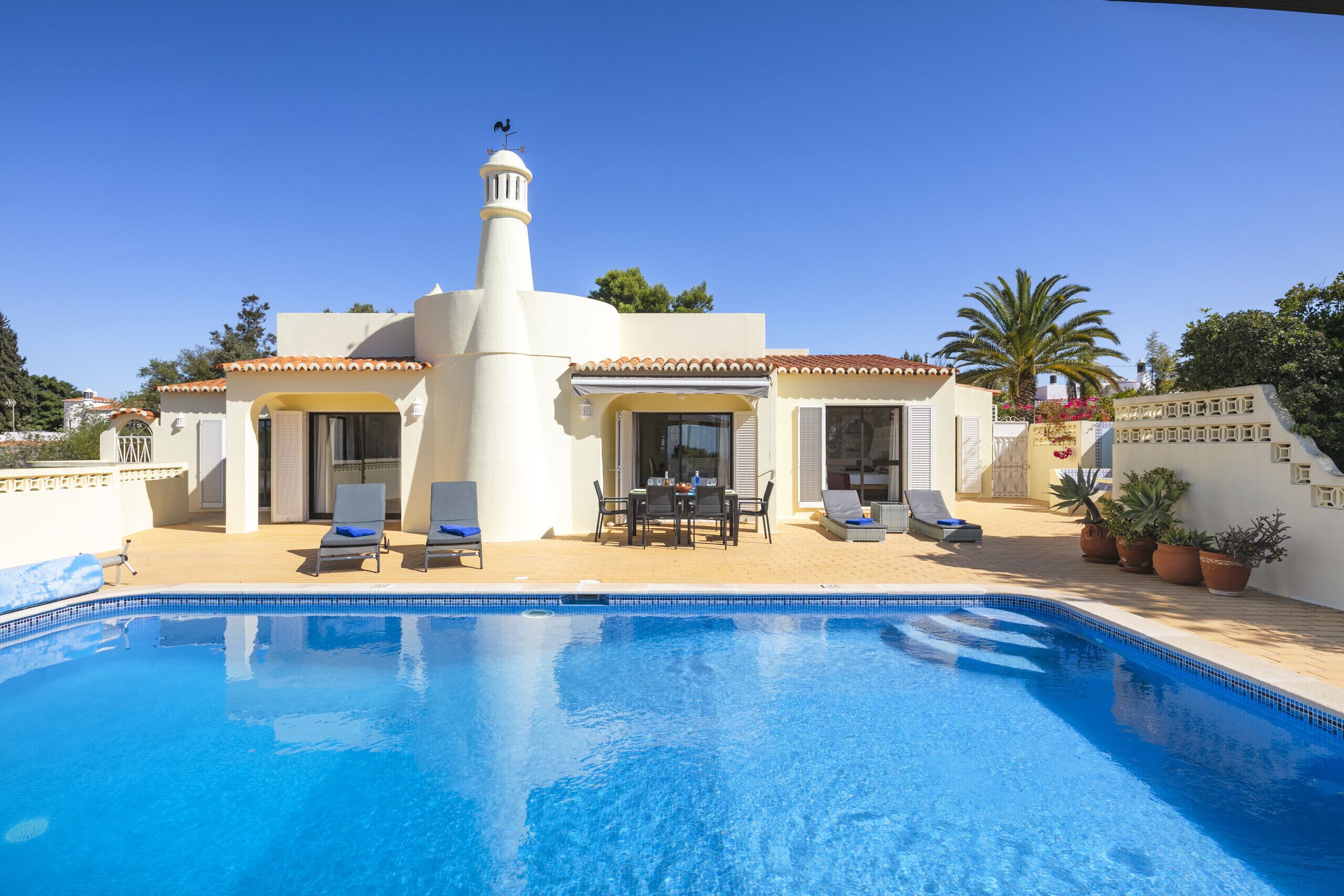 A poolside area with a clear blue pool, lounge chairs, and a white building with a tower.