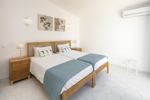A double bed with a wooden frame and a blue blanket, a wooden nightstand with a lamp, and a small white stool.